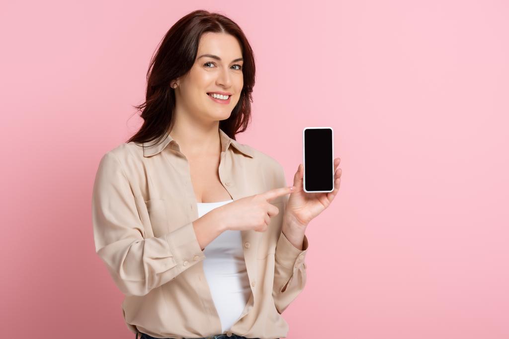 Hymyilevä nainen, joka osoittaa sormella älypuhelinta vaaleanpunaisella taustalla, kehon positiivinen käsite  - Valokuva, kuva