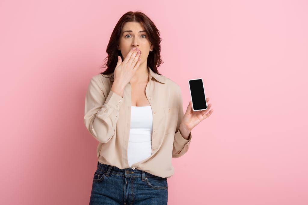 Σοκαρισμένη γυναίκα που καλύπτει το στόμα με το χέρι και δείχνει smartphone σε ροζ φόντο, έννοια του σώματος θετική  - Φωτογραφία, εικόνα