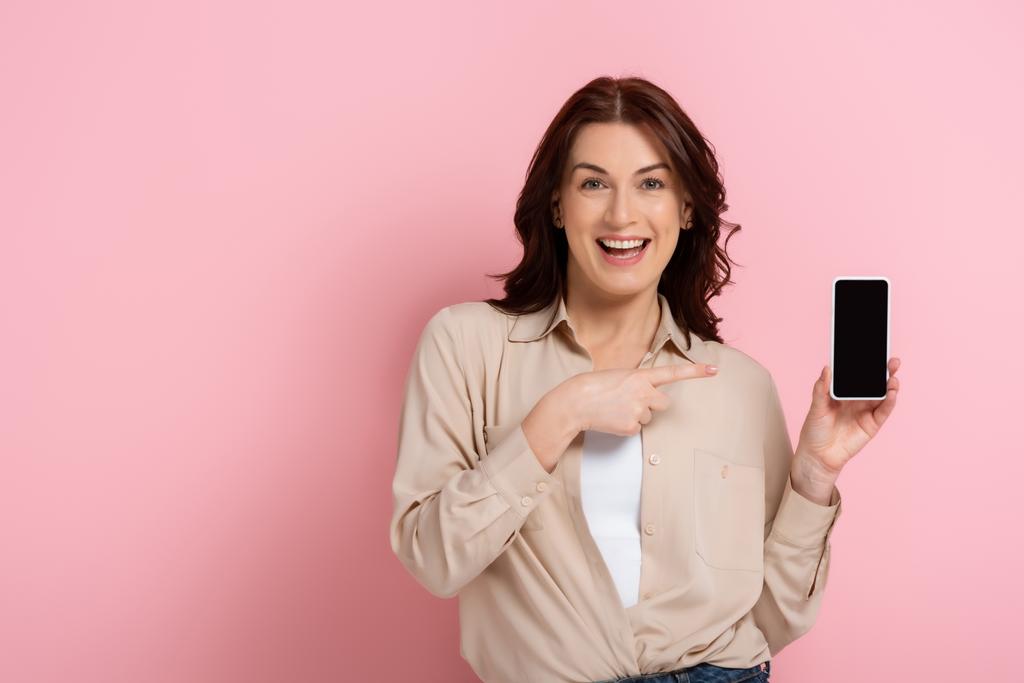 Schöne Frau lächelt in die Kamera und zeigt mit dem Finger auf Smartphone mit leerem Bildschirm auf rosa Hintergrund - Foto, Bild
