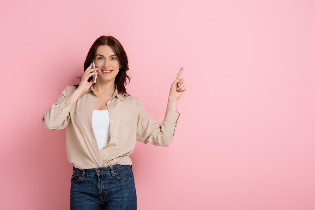 Beautiful brunette woman pointing with finger while talking on smartphone on pink background - Photo, Image