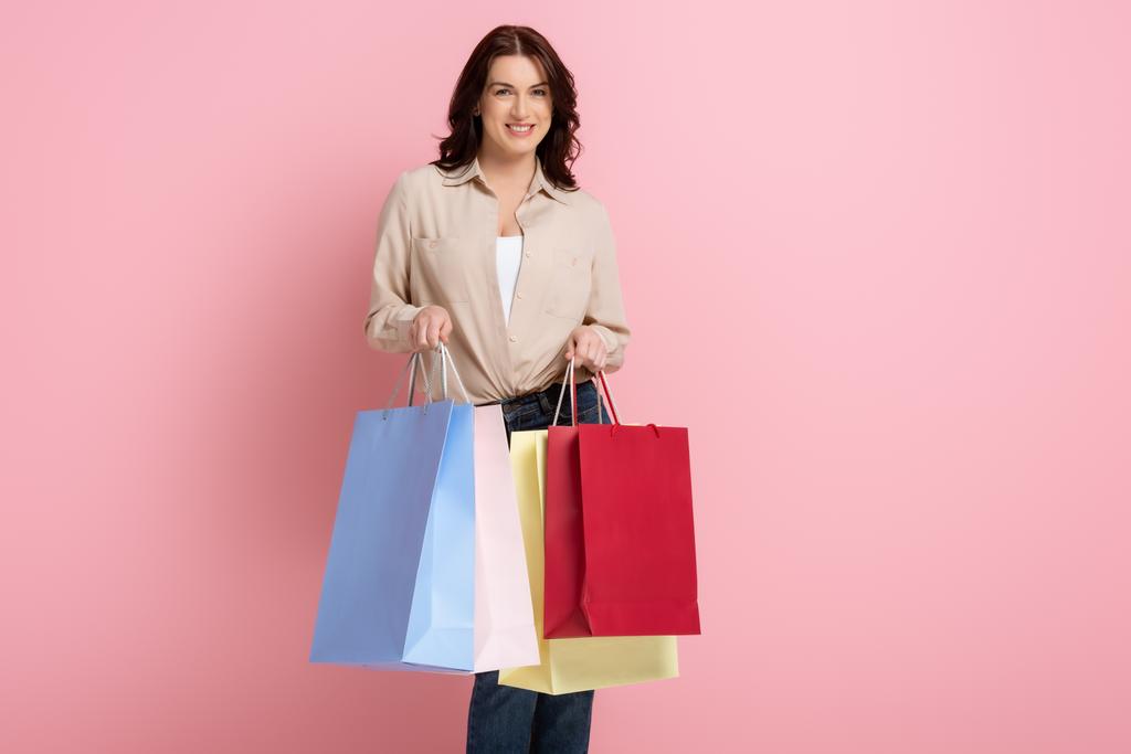 Hermosa mujer sonriendo mientras sostiene coloridas bolsas de compras sobre fondo rosa
 - Foto, Imagen