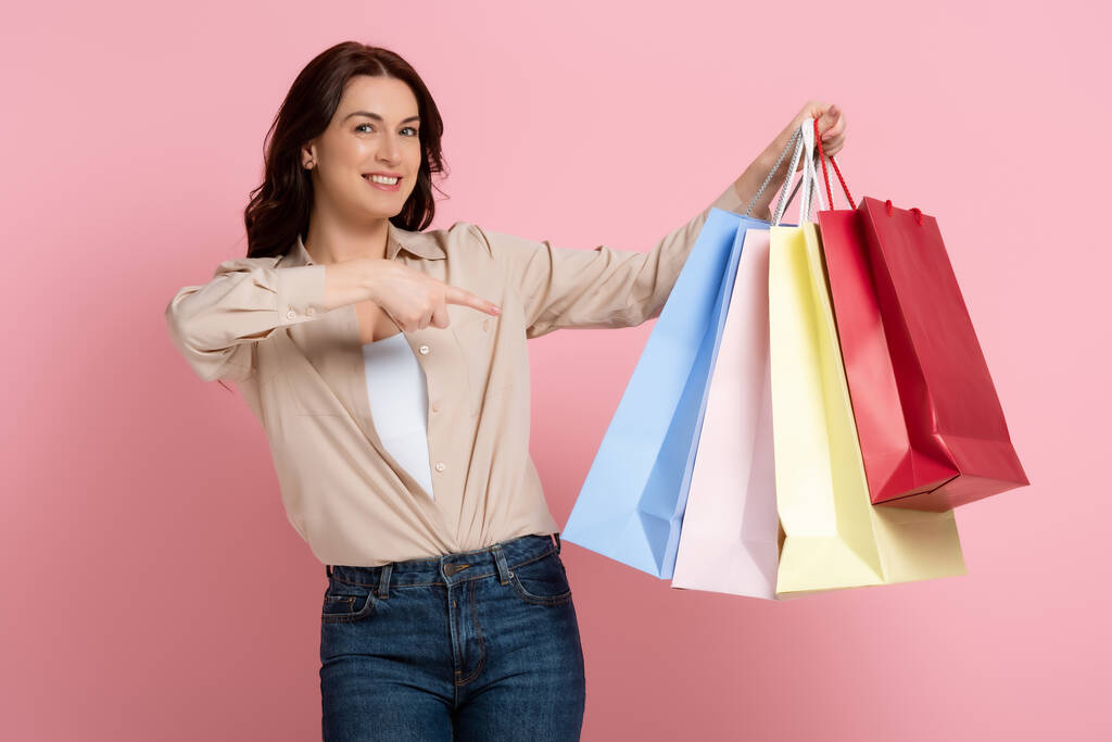 Beautiful brunette woman smiling at camera and pointing with finger at colorful shopping bags on pink background - Photo, Image