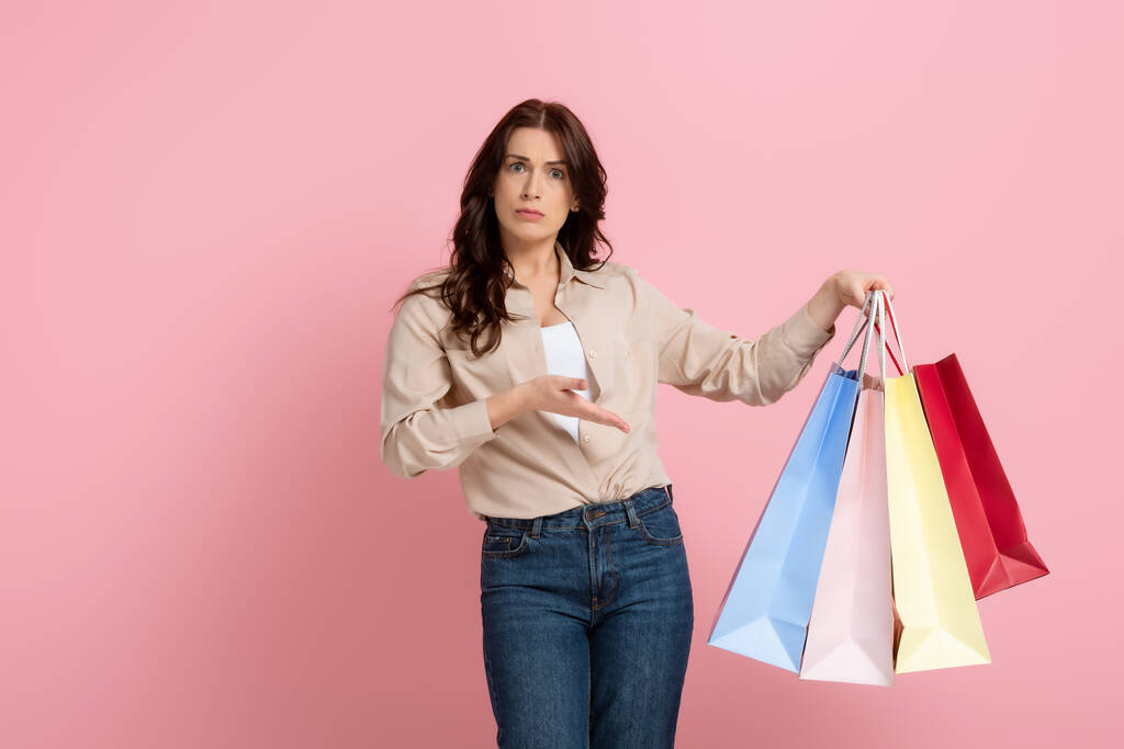 Skeptische Frau zeigt mit der Hand auf bunte Einkaufstüten auf rosa Hintergrund - Foto, Bild
