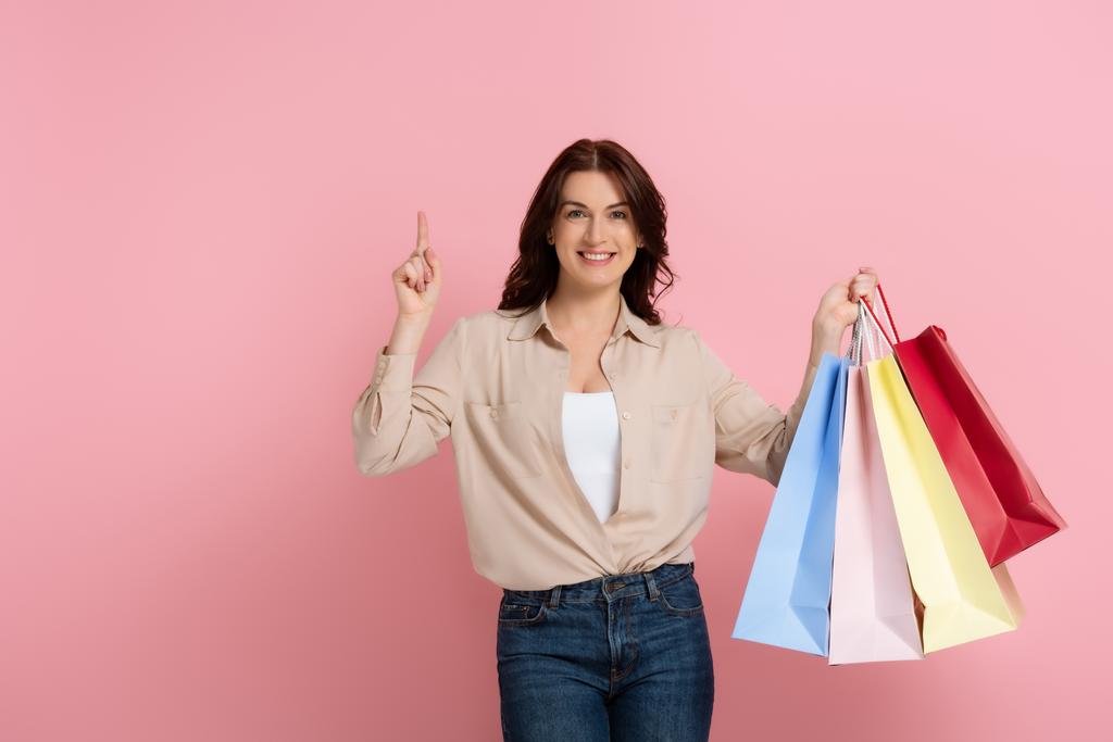 Positive brünette Frau zeigt mit dem Finger, während sie Einkaufstüten auf rosa Hintergrund hält - Foto, Bild