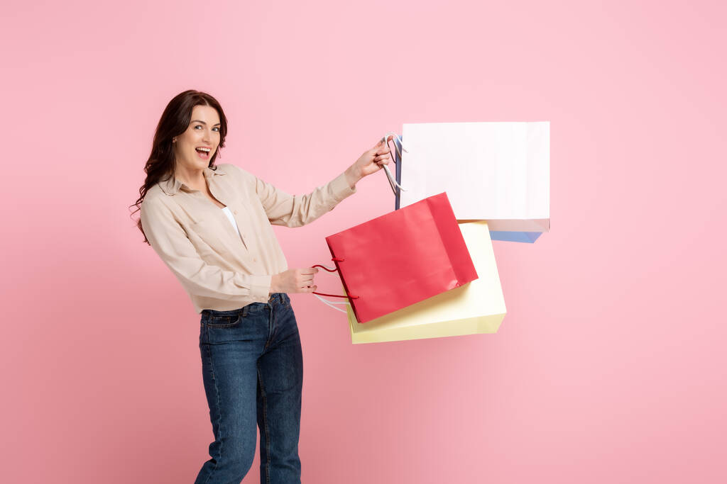 Femme joyeuse regardant la caméra tout en tenant des sacs à provisions colorés isolés sur rose, concept de corps positif  - Photo, image