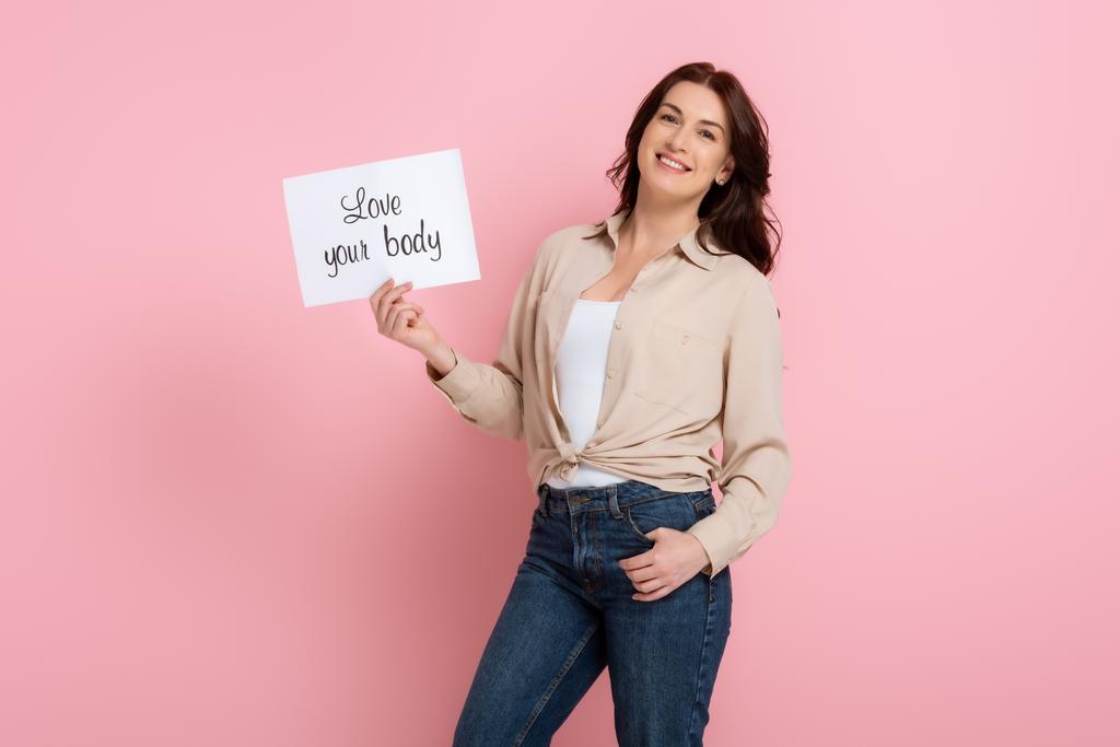 Усміхнена жінка тримає листівку з любов'ю, що витісняє ваше тіло на рожевому фоні, концепція позитивного тіла
 - Фото, зображення