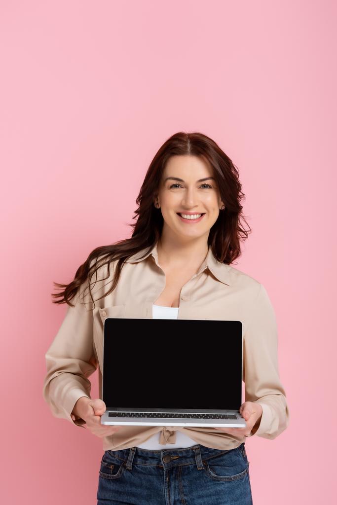 Όμορφη χαμογελαστή γυναίκα δείχνει φορητό υπολογιστή με κενή οθόνη απομονώνονται σε ροζ  - Φωτογραφία, εικόνα