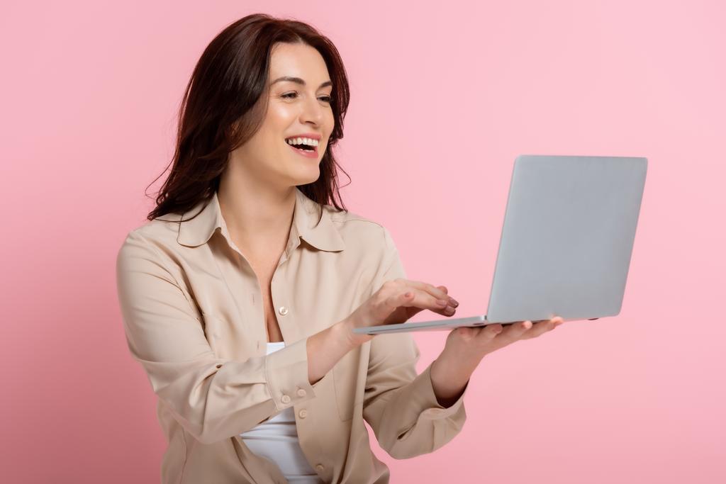 Ελκυστική μελαχρινή γυναίκα χαμογελώντας κατά τη χρήση φορητού υπολογιστή σε ροζ φόντο - Φωτογραφία, εικόνα