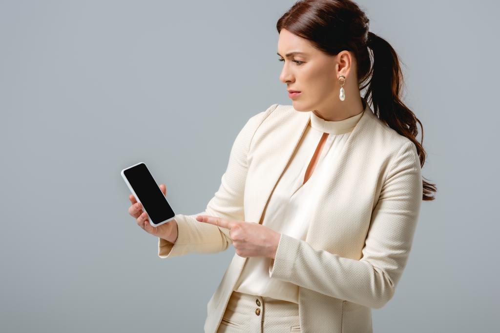 Mooie vrouw in formele kleding wijzend met de vinger naar smartphone geïsoleerd op grijs  - Foto, afbeelding