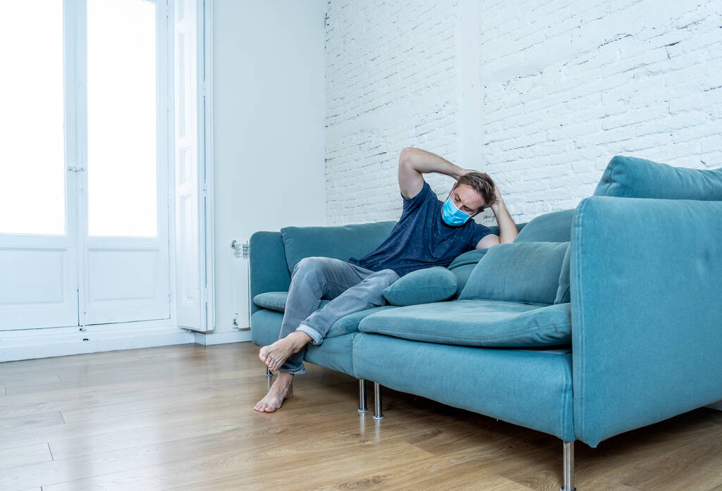 Грустный мужчина в защитной маске на домашнем диване чувствует усталость и беспокойство, страдая депрессией в условиях блокировки коронавируса и социальной дистанцированности. Концепция психического здоровья и изоляции
. - Фото, изображение