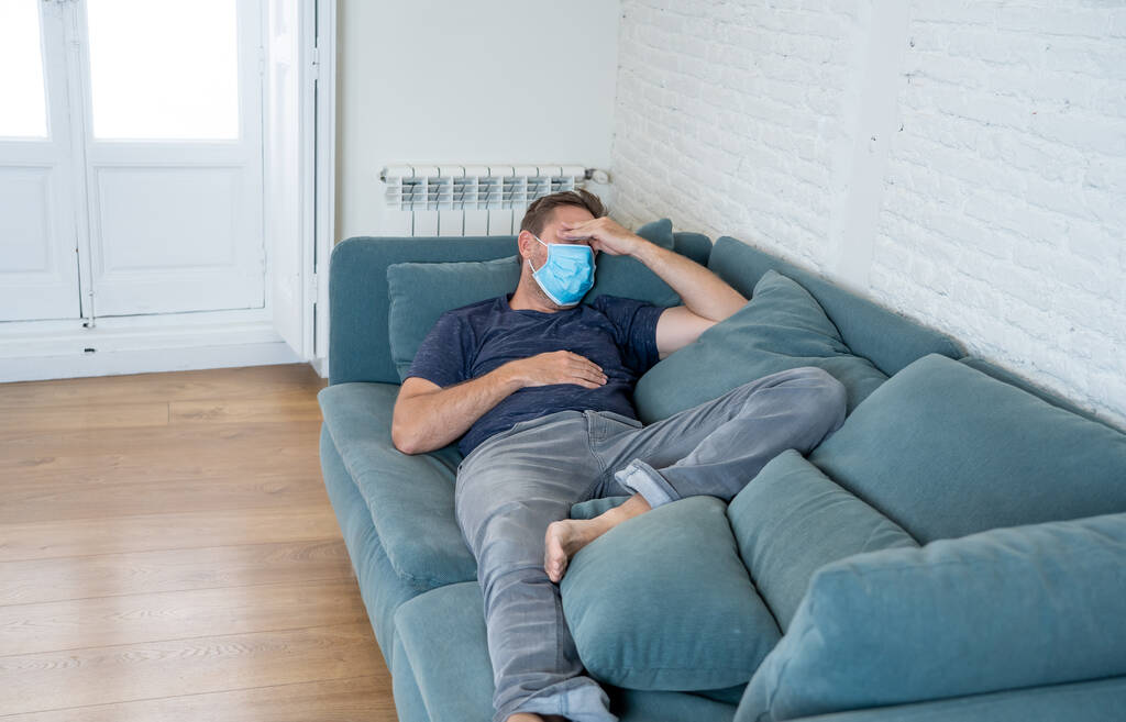 自宅のリビングルームのソファで保護顔マスクを持つ悲しい男は、疲れやコロナウイルスのロックダウンや社会的距離の中での苦しみうつ病を心配感じています。精神衛生と隔離の概念. - 写真・画像