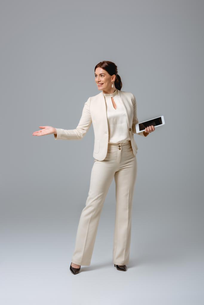 Smiling businesswoman pointing with hand and holding digital tablet on grey background - Photo, Image