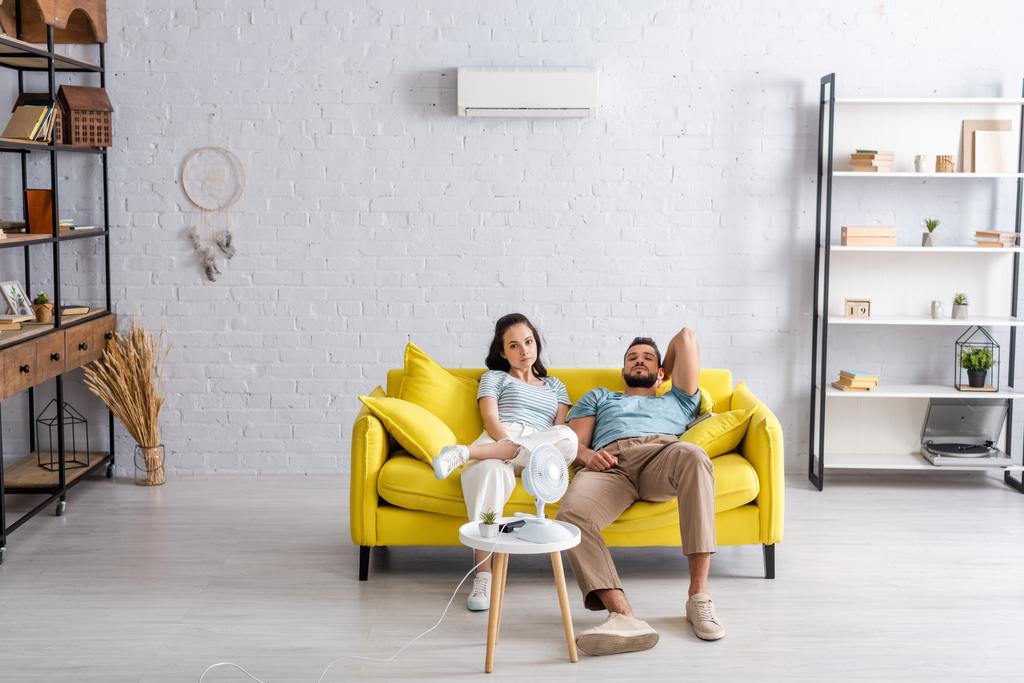 Молода пара дивиться на камеру, сидячи на дивані біля електричного вентилятора і кондиціонера на стіні у вітальні
 - Фото, зображення