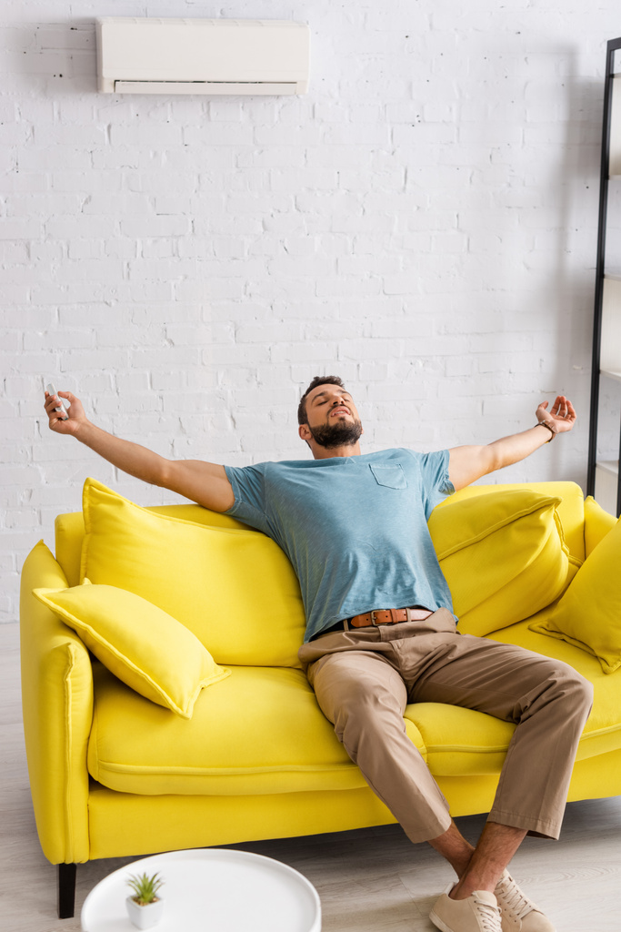 Fiatal férfi kezében távirányító a légkondicionáló, miközben ül csukott szemmel kanapén otthon  - Fotó, kép