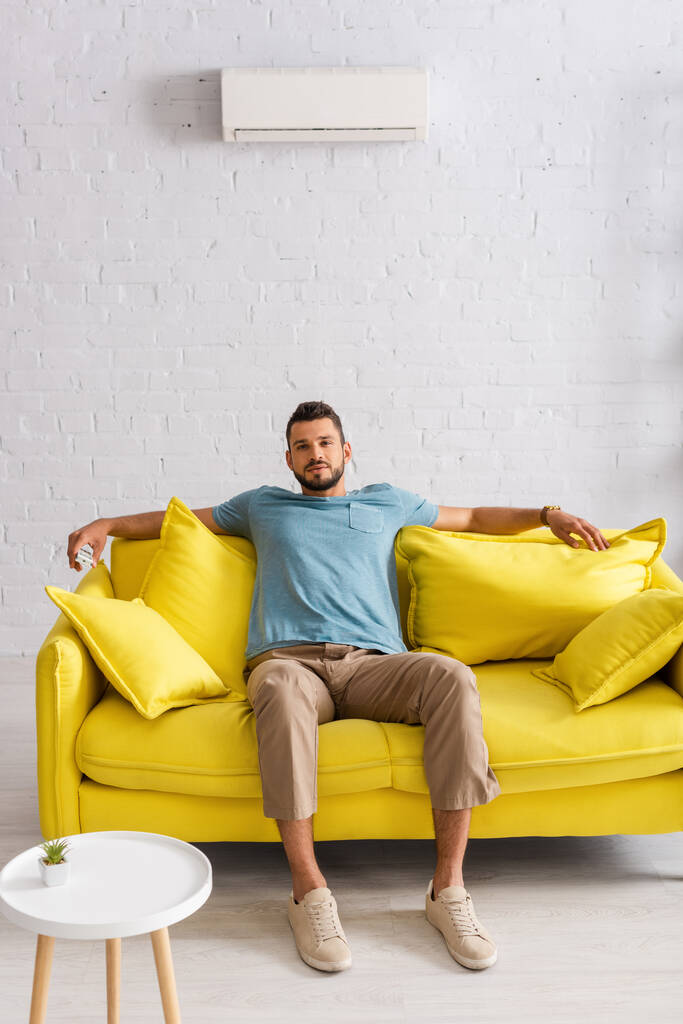 Valikoiva painopiste komea mies tilalla kaukosäädin ilmastointilaite katsoessaan kameran sohvalla kotona  - Valokuva, kuva