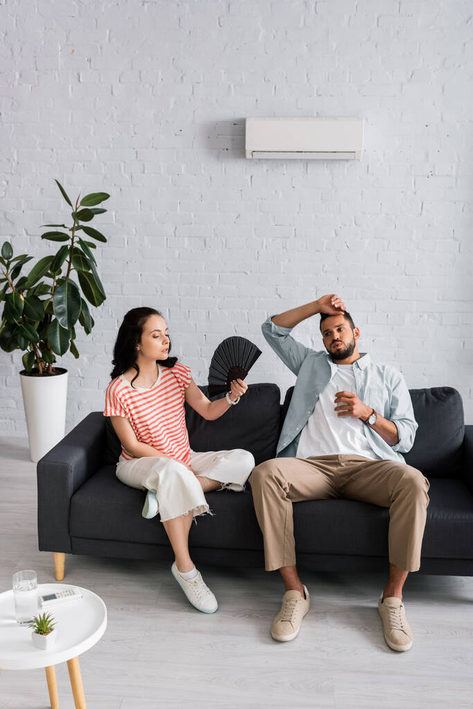 Erschöpfter Mann hält Wasserglas neben Freundin mit Ventilator auf Couch  - Foto, Bild