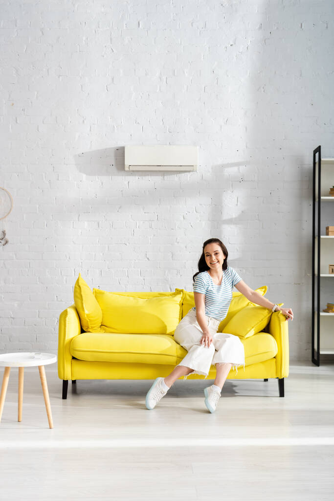 Houkutteleva tyttö hymyilee kameralle istuessaan sohvalla lähellä kauko-ohjain ilmastointilaite sohvapöytä kotona  - Valokuva, kuva