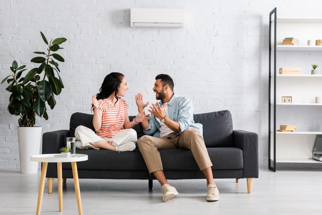 Positive couple smiling at each other while sitting on sofa under air conditioner at home  - Photo, Image