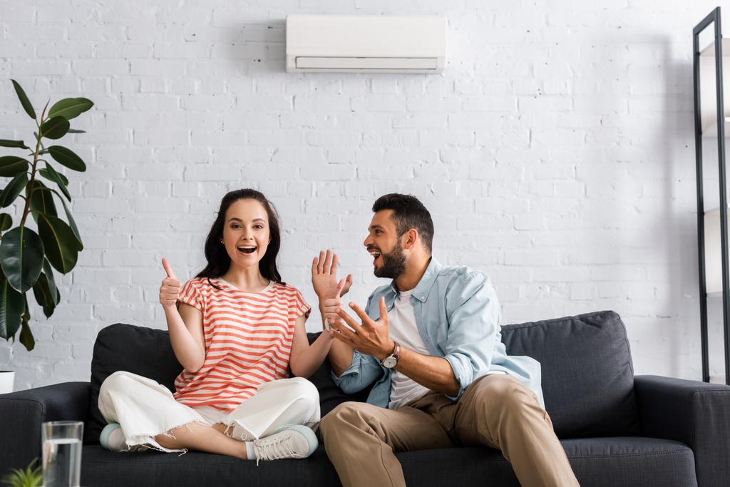 Foco seletivo de mulher sorridente mostrando polegares para cima perto de namorado alegre na sala de estar
 - Foto, Imagem