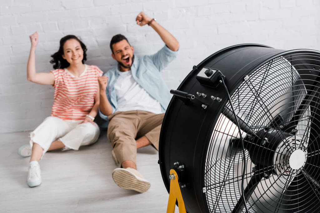 Selective focus of cheerful couple showing yeah gesture while sitting near electric fan on floor  - Photo, Image