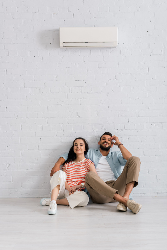 Jeune couple souriant à la caméra assis sous le climatiseur sur le sol à la maison - Photo, image