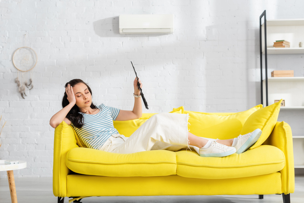Selektiver Fokus eines müden Mädchens, das einen Ventilator hält, während es auf dem Sofa im Wohnzimmer unter Hitze leidet - Foto, Bild
