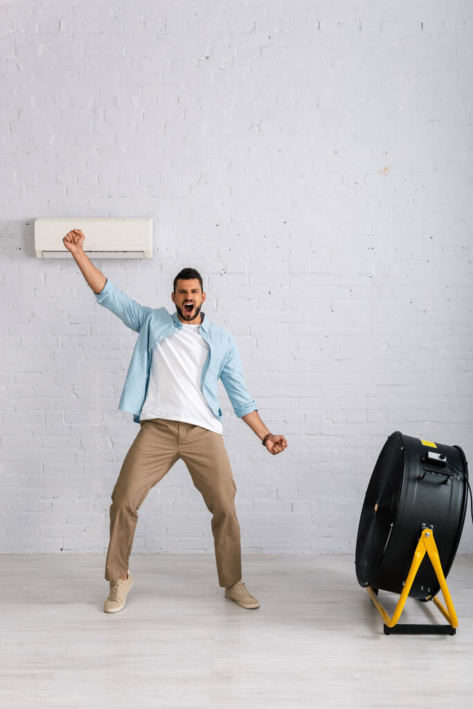 Pozytywny mężczyzna pokazuje tak gest stojąc w pobliżu wentylatora elektrycznego i klimatyzatora w domu  - Zdjęcie, obraz