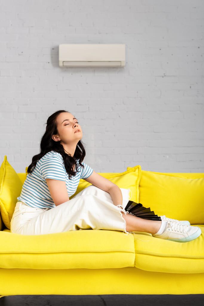 Vista lateral da menina exausta segurando ventilador enquanto sentado no sofá na sala de estar
 - Foto, Imagem