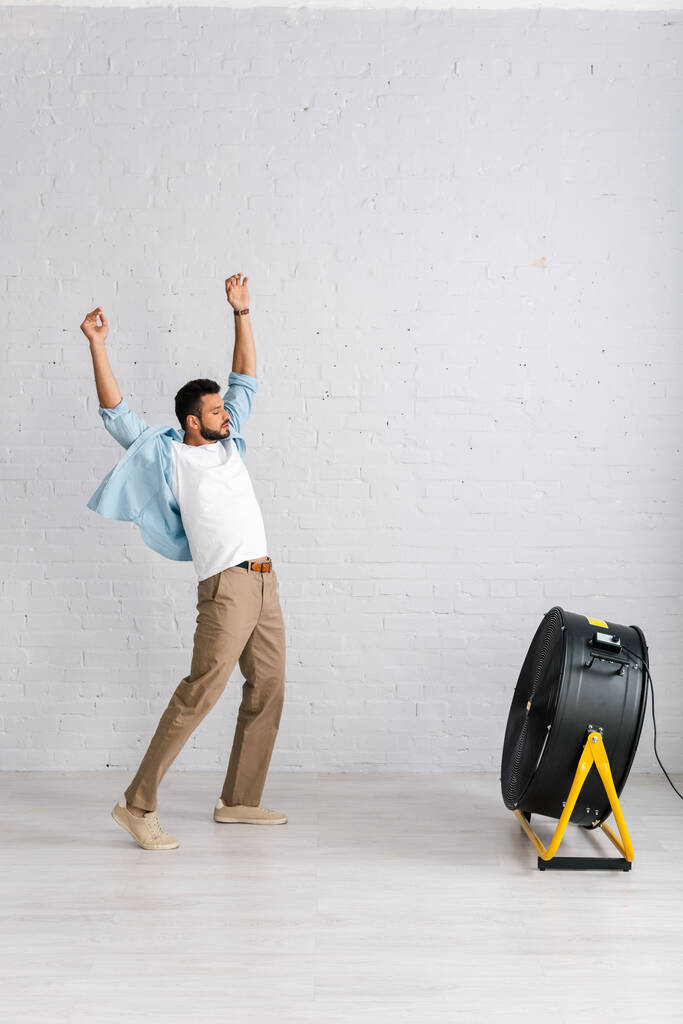 Hezký vousatý muž stojící u elektrického ventilátoru v obývacím pokoji - Fotografie, Obrázek