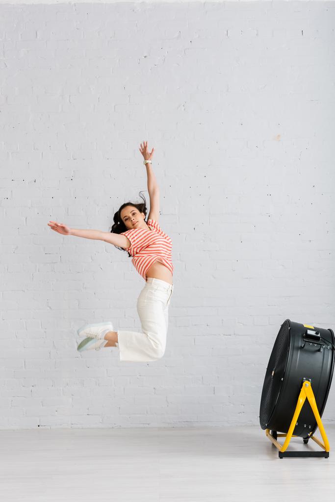 Jonge vrouw springen met opgeheven handen in de buurt van elektrische ventilator op vloer thuis - Foto, afbeelding