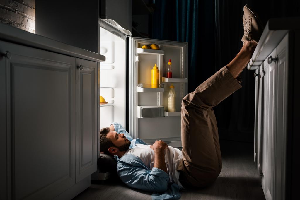 Vista laterale o uomo che dorme al piano vicino al frigorifero aperto di notte  - Foto, immagini