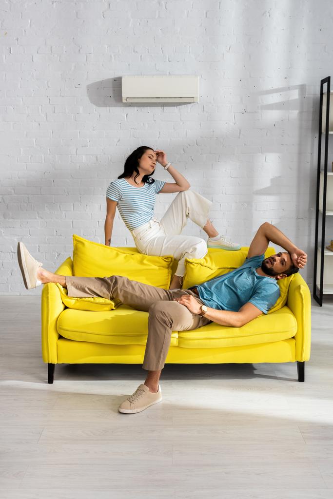 リビングルームでのバックグラウンドで壁にエアコン付きのソファに座っている間、若いカップルは熱い感じ - 写真・画像