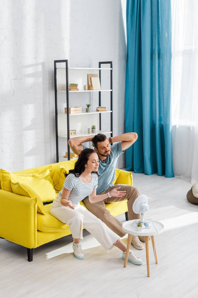 Young couple sitting on couch near electric fan on coffee table in living room - Photo, Image