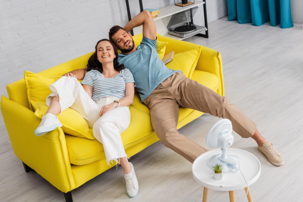 Enfoque selectivo de pareja positiva acostada en el sofá con los ojos cerrados cerca de ventilador eléctrico en la mesa de café
  - Foto, Imagen