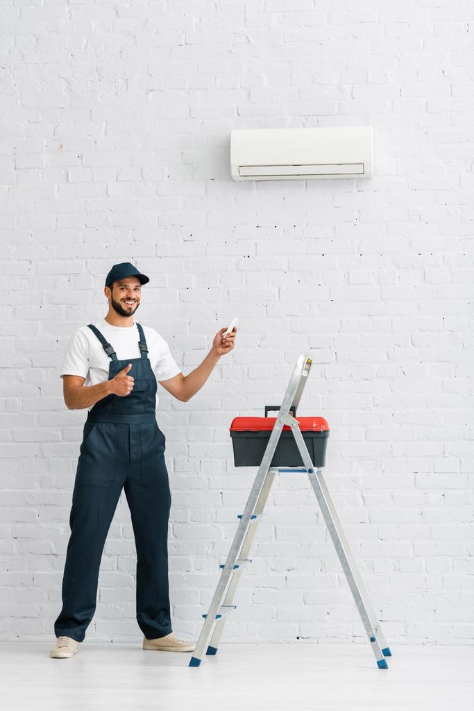 Trabajador sonriente mostrando el pulgar hacia arriba mientras sostiene el controlador remoto del aire acondicionado cerca de la caja de herramientas en la escalera
  - Foto, imagen