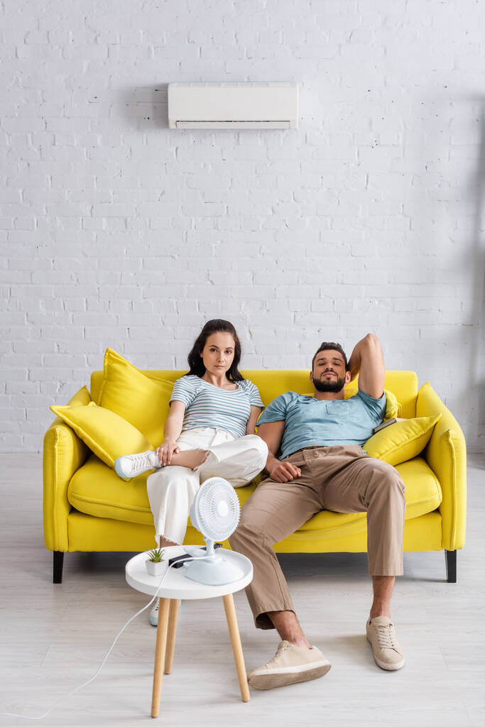 Enfoque selectivo de pareja joven mirando la cámara cerca del ventilador eléctrico en la mesa de café en la sala de estar
 - Foto, Imagen