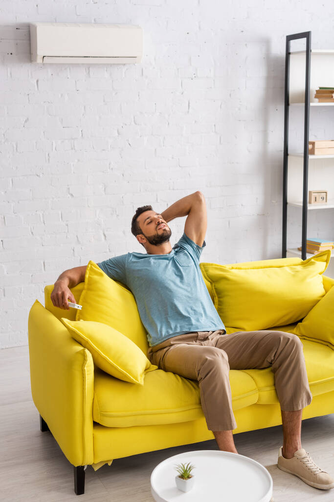 Hombre guapo sosteniendo mando a distancia de aire acondicionado en el sofá en la sala de estar
 - Foto, imagen