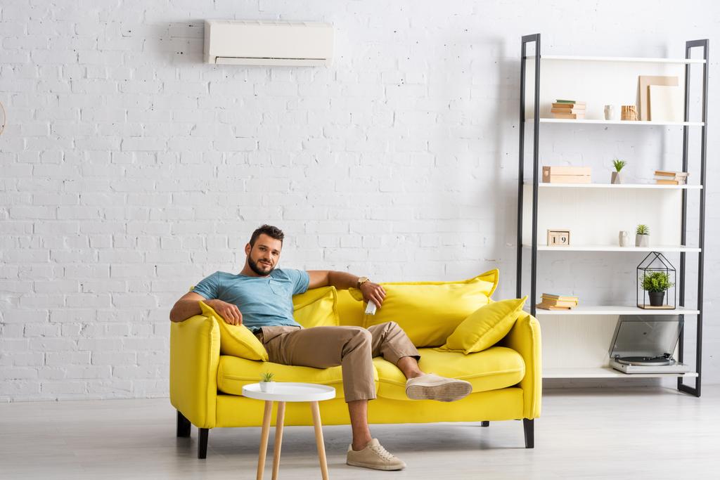 Hombre barbudo guapo mirando a la cámara mientras sostiene el mando a distancia del aire acondicionado en la sala de estar
 - Foto, imagen