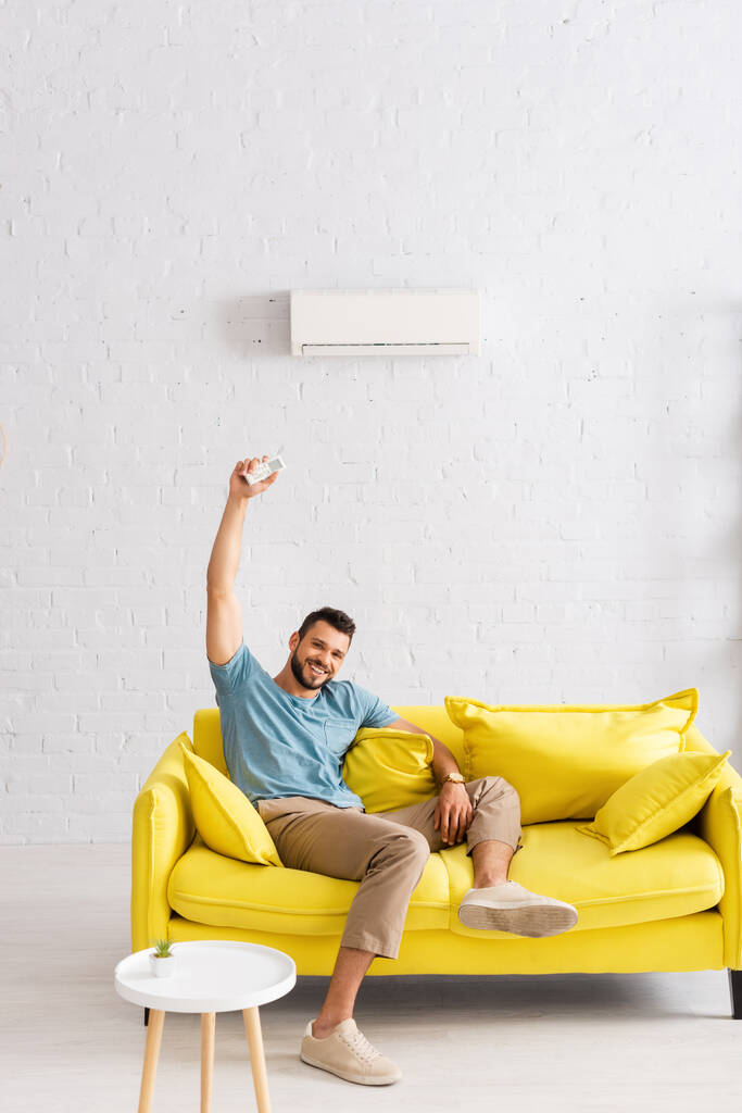 Vrolijke man lacht naar de camera terwijl hij de afstandsbediening van de airconditioner in de woonkamer vasthoudt - Foto, afbeelding