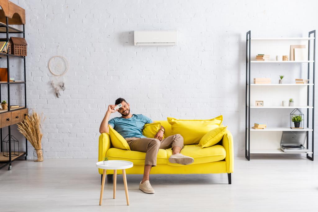 Positiivinen mies, jolla ilmastointilaitteen kaukosäädin ja hymyilevä kamera sohvalla kotona  - Valokuva, kuva