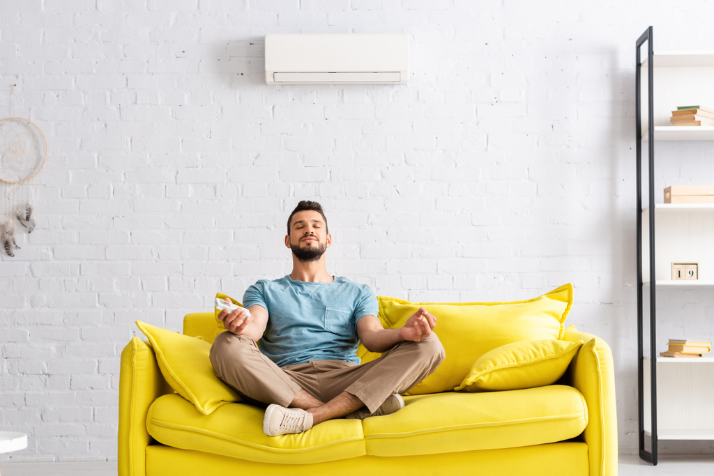 Fiatal férfi kezében távirányító a légkondicionáló, miközben meditál kanapén  - Fotó, kép