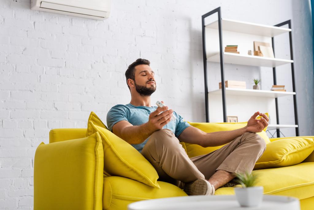 Enfoque selectivo del hombre guapo meditando mientras sostiene el mando a distancia del aire acondicionado en el sofá
  - Foto, imagen