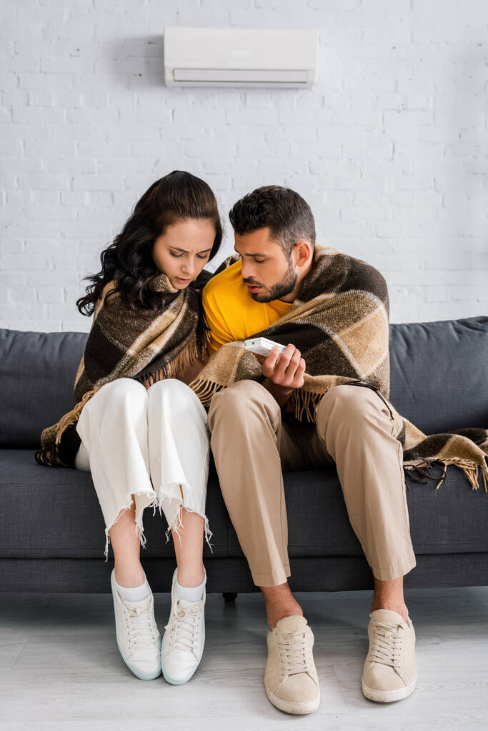Jeune homme tenant télécommande du climatiseur tout en se sentant froid près de la petite amie sur le canapé à la maison  - Photo, image