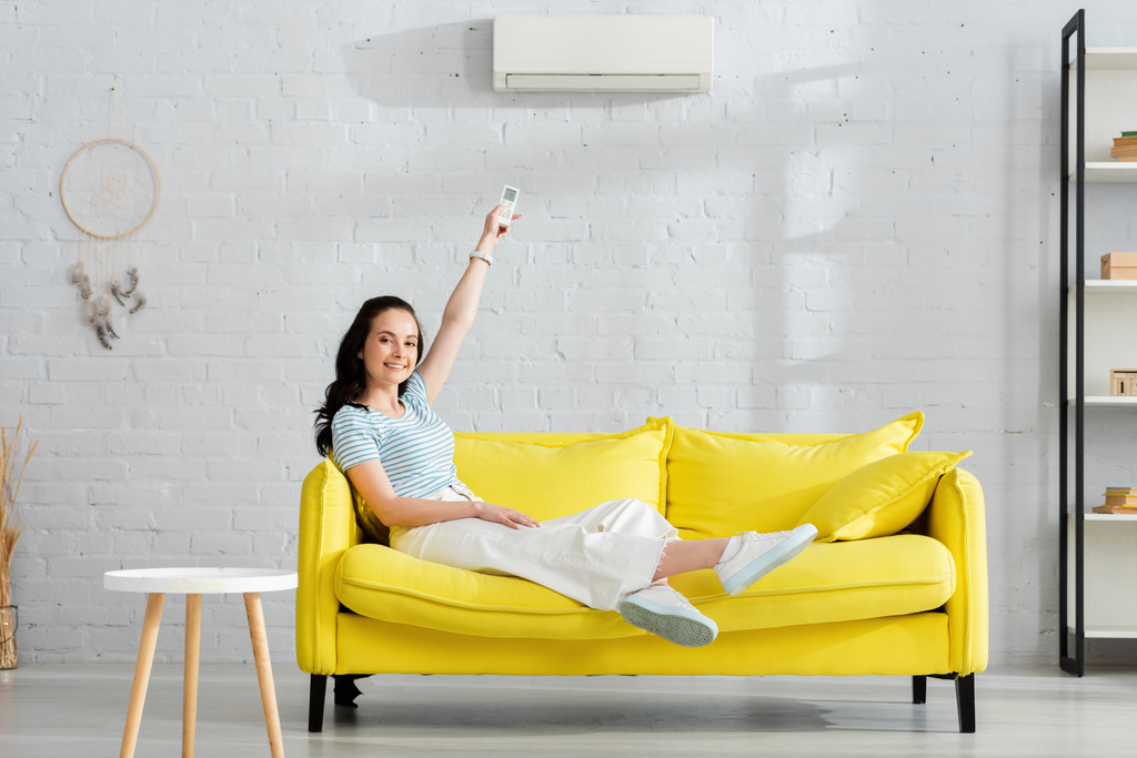 Όμορφη κοπέλα χαμογελώντας στην κάμερα, ενώ εναλλαγή κλιματιστικού με τηλεχειριστήριο στο σπίτι  - Φωτογραφία, εικόνα