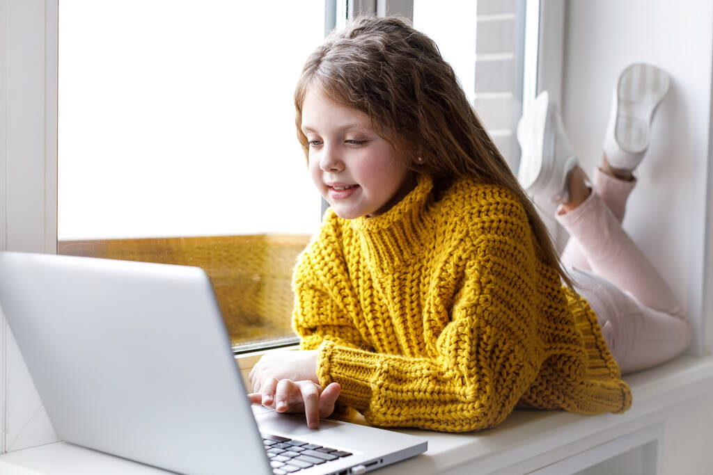 Ένα όμορφο κορίτσι με ένα φορητό υπολογιστή στο σπίτι στο παράθυρο κοιτάζει την οθόνη και χαμογελά. Το κορίτσι στο σπίτι επικοινωνεί στο διαδίκτυο. Παιδιά εξ αποστάσεως εκπαίδευση. - Φωτογραφία, εικόνα