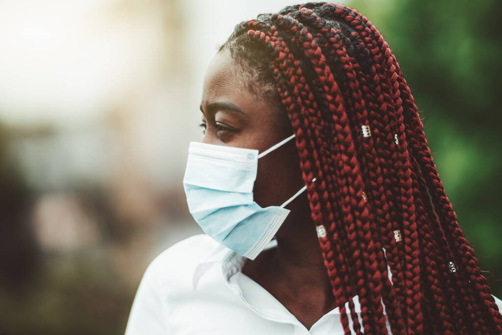 Retrato de close-up de uma jovem africana com tranças de castanha e com uma máscara protetora do vírus sobre o rosto; mulher negra mascarada ao ar livre - proteção contra a gripe e o foco seletivo e pandêmico
 - Foto, Imagem
