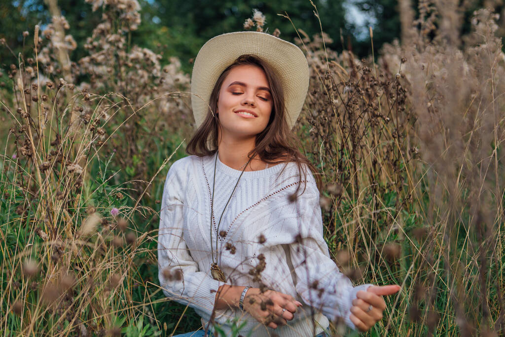 美しいブルネットの十代の女の子は白いセーター、ジーンズとカウボーイストローの帽子を身に着けています乾燥した茶色のバーの草の中に座って、笑顔と日没の間に笑って - 写真・画像