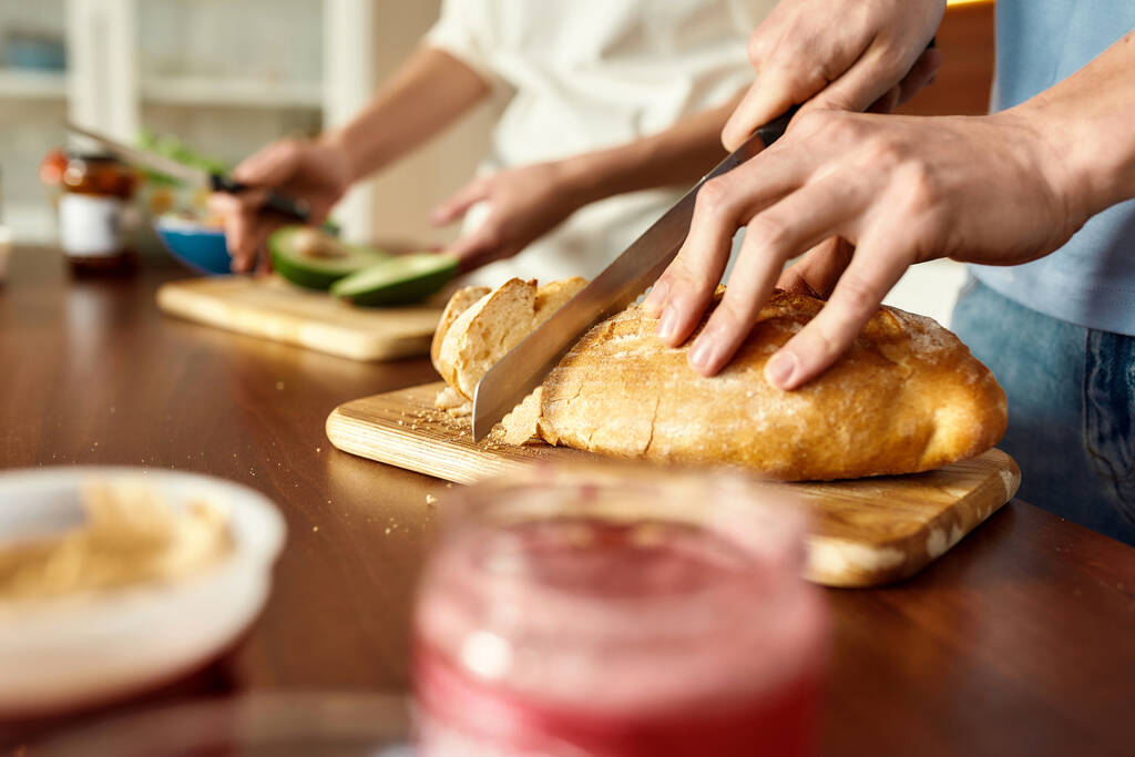Αρσενικά χέρια κόβουν νόστιμο σπιτικό ψωμί στον ξύλινο πίνακα. Νεαρό ζευγάρι ετοιμάζεται υγιεινό γεύμα στην κουζίνα μαζί. Χορτοφαγία, υγιεινή διατροφή έννοια. - Φωτογραφία, εικόνα