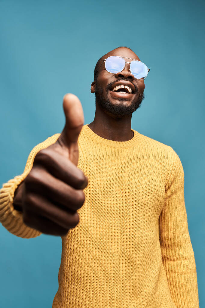 retrato foto de un chico guapo de piel oscura con una barba sobre un fondo azul en un suéter amarillo, gafas transparentes, mira a la cámara, sonríe y mantiene su pulgar hacia arriba
 - Foto, Imagen