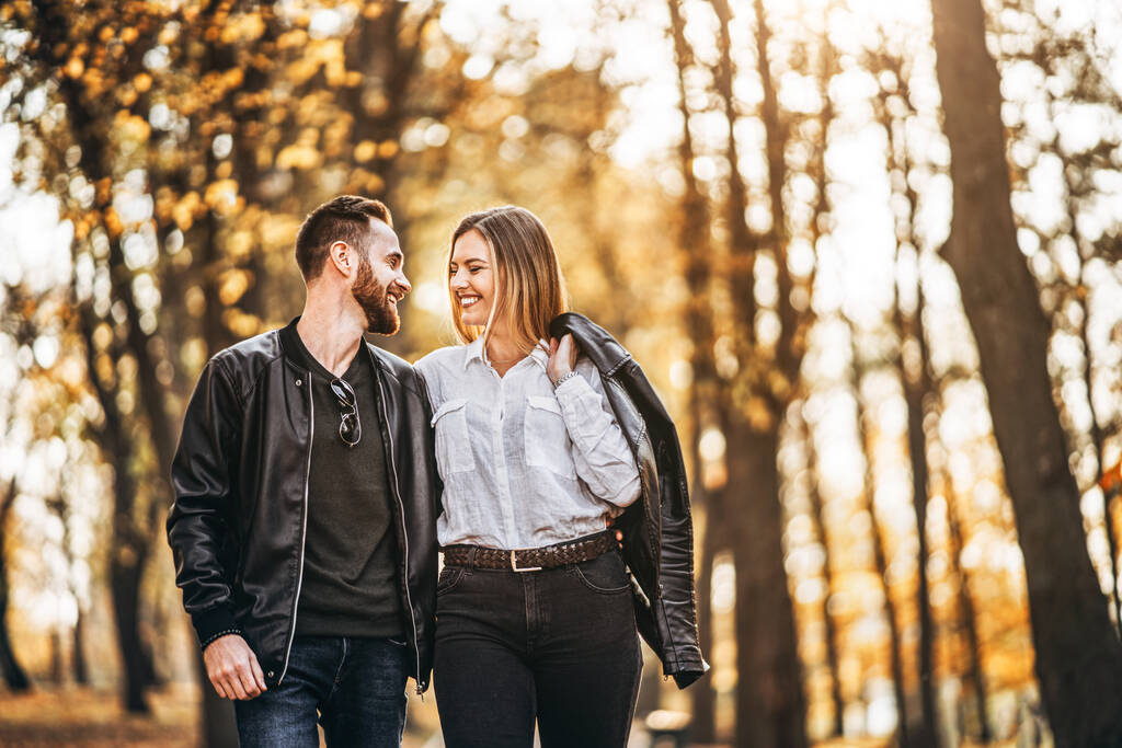 Πορτρέτο ενός ευτυχισμένου ζευγαριού που περπατάει στο πάρκο του φθινοπώρου. Άντρας και γυναίκα κρατιούνται χέρι-χέρι και χαμογελούν. - Φωτογραφία, εικόνα